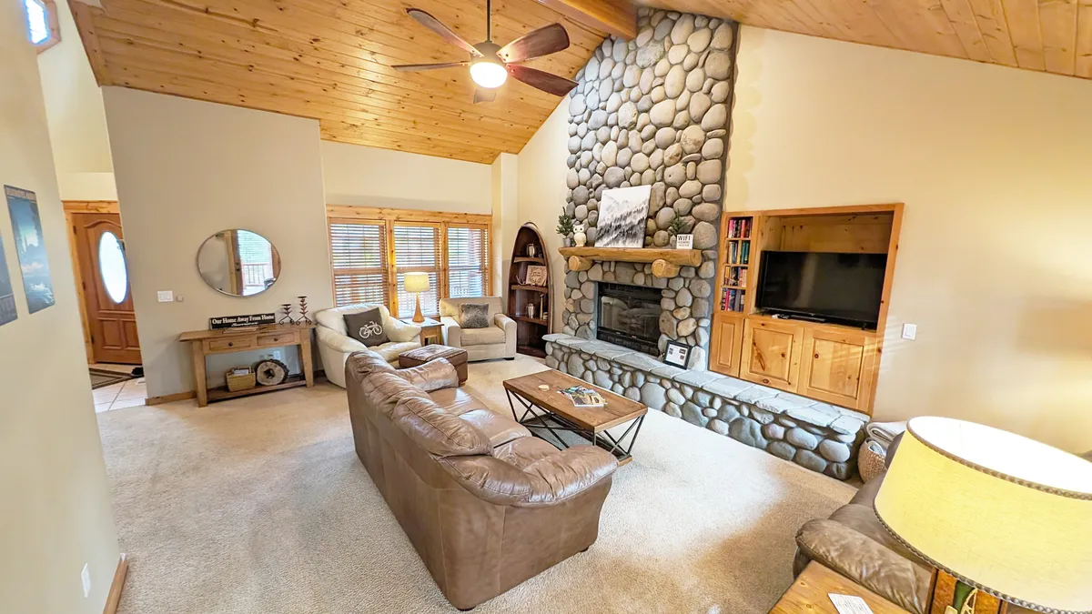 A cozy living room featuring a stone fireplace as the focal point, surrounded by a large leather couch, a wooden coffee table, and expansive windows that let natural light pour in. The space feels warm and inviting, perfect for gatherings and relaxation.