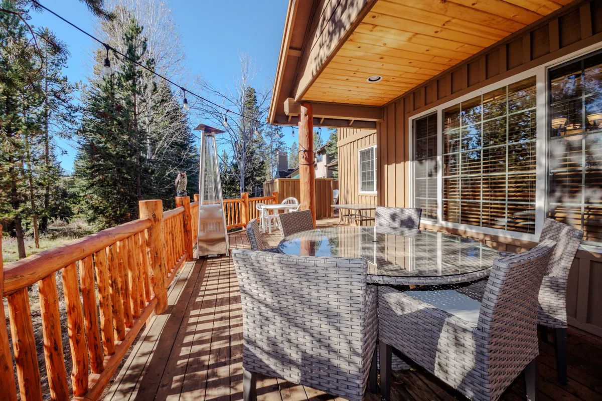 A bright, friendly patio scene features a glass-top table with woven chairs, a patio heater, and natural wood railings. The lush forest backdrop sets the tone for relaxed outdoor meals, morning coffees, or celebratory gatherings with family and friends.