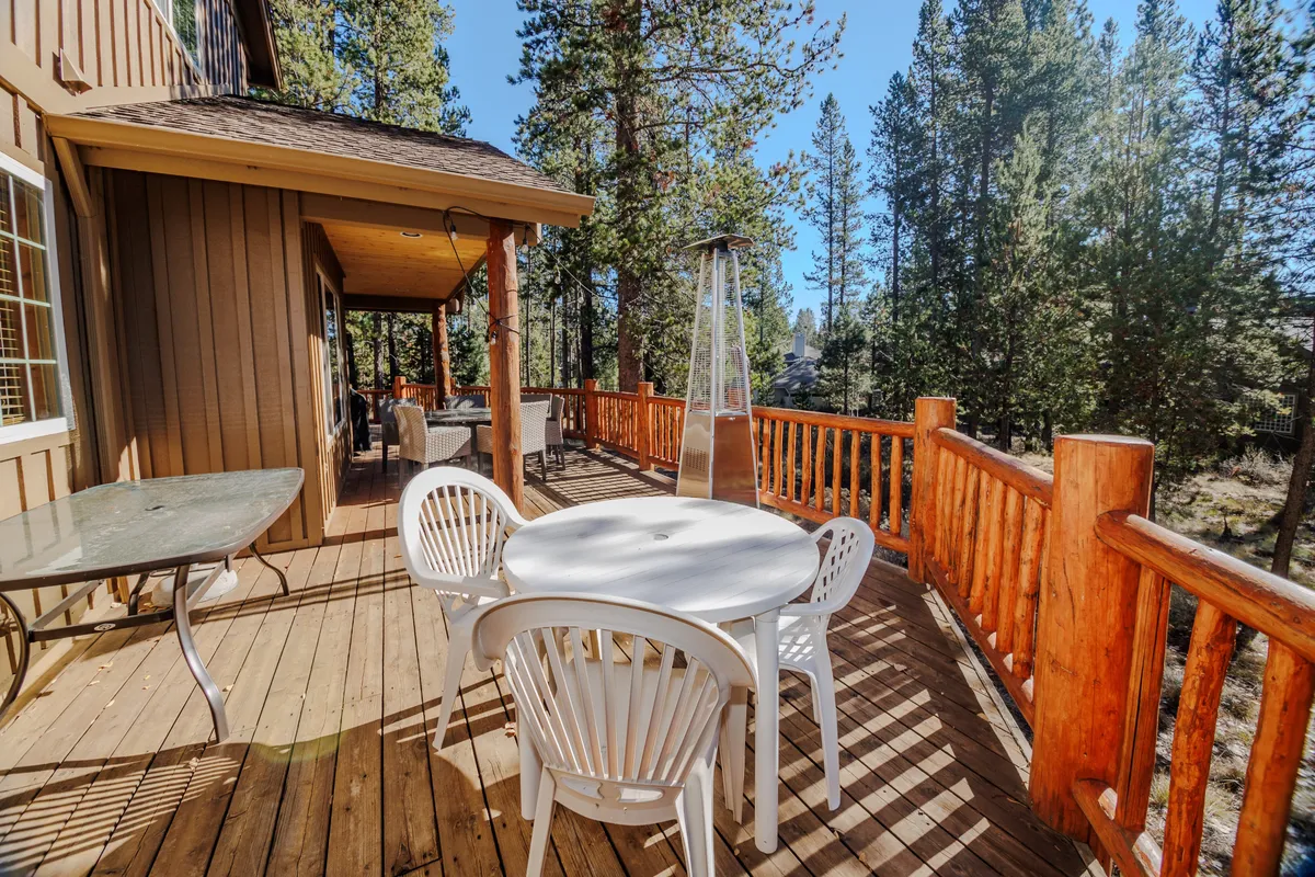 An inviting outdoor dining area with a glass table and woven chairs sits atop a wooden deck, surrounded by a patio heater and natural wooden railing. Tall trees and greenery envelop the space, creating a tranquil setting for meals and gatherings in the fresh mountain air.