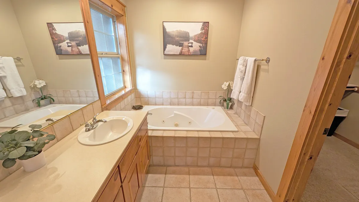 A luxurious bathroom featuring a jacuzzi tub, expansive mirror, window letting in natural light, and a sleek sink area. Framed artwork and inviting finishes contribute to a tranquil, spa-like retreat ideal for unwinding after adventurous days.