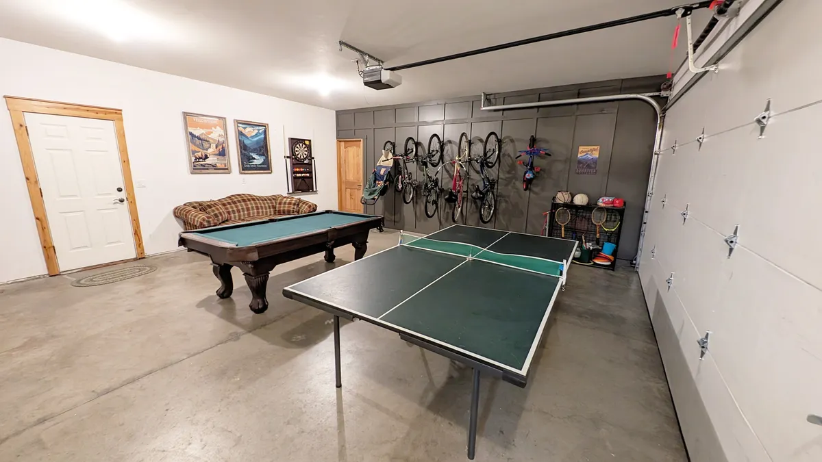 A lively recreation garage featuring a pool table and ping pong table, with a cozy couch for spectators, visible dartboard, bicycle storage, and playful wall art. The space is open, well-lit, and clearly designed for active family fun, group games, and friendly competition.