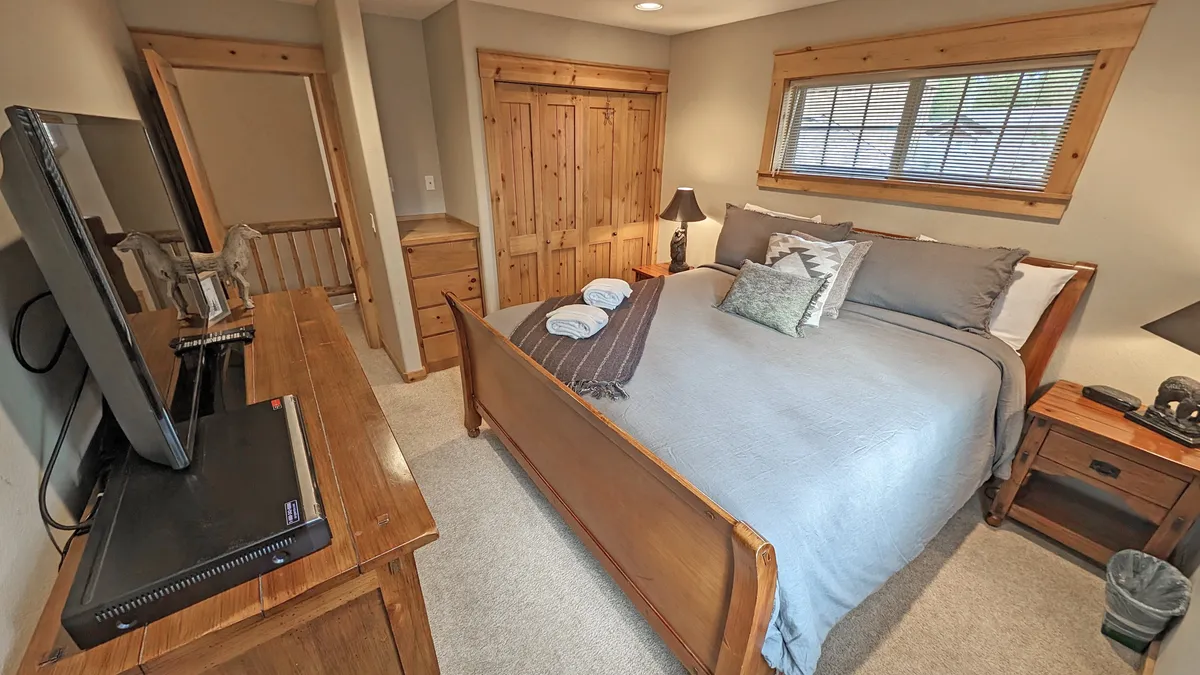 This image features a king bedroom with a neatly made bed dressed in crisp linens and plush pillows. Wooden furniture, including a dresser and nightstands, adds warmth and functionality, while a television sits atop the dresser for in-room entertainment. Natural light filters in, creating a restful and inviting atmosphere.