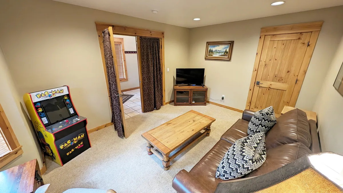 A warm, inviting den setup centered on a plush brown sofa, a wooden coffee table, a TV on a wooden stand, and a mini Pac-Man arcade machine glowing in the corner. The ambiance hints at both classic and modern gaming, perfect for movie nights or a quiet spot for board games.