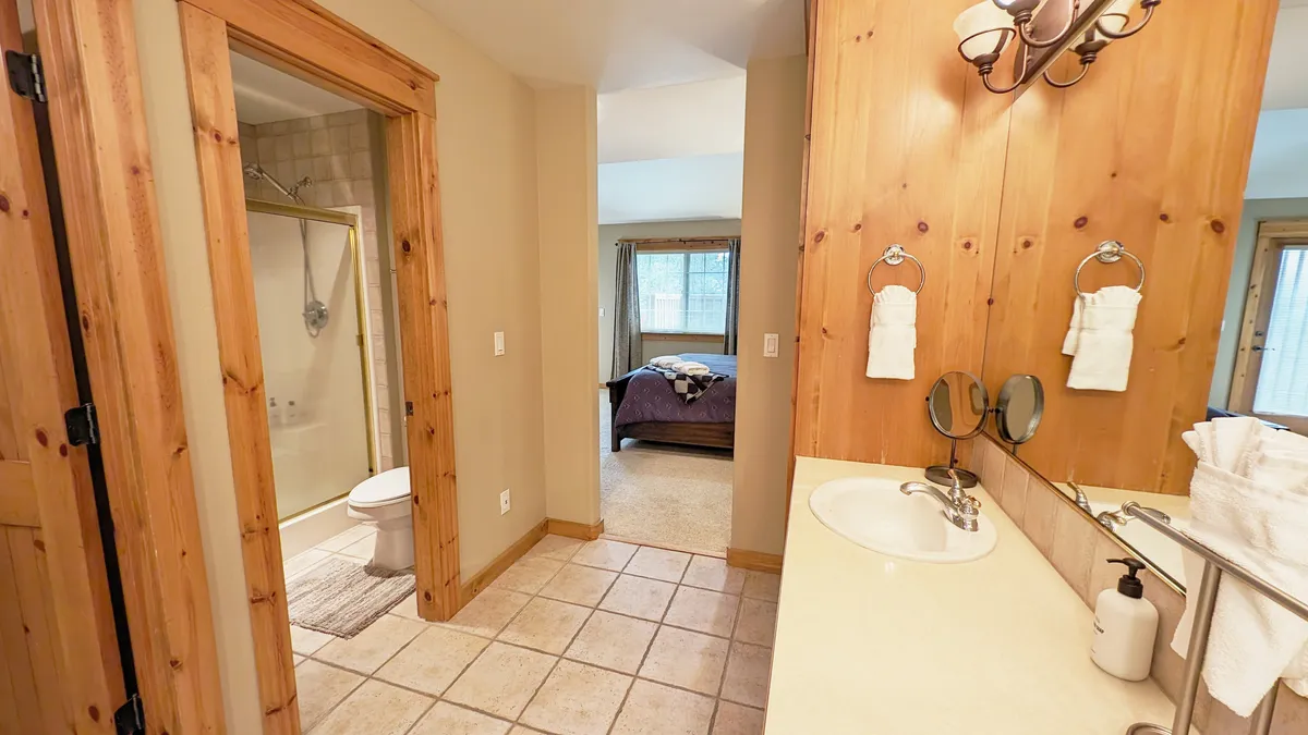 A serene bathroom scene with warm wooden paneling, a large mirror, and a vanity with a spacious sink. A cozy glimpse of a bedroom through an open doorway adds context. The space is designed for relaxation and personal grooming, with soft morning light streaming through the window, and amenities that support daily routines.